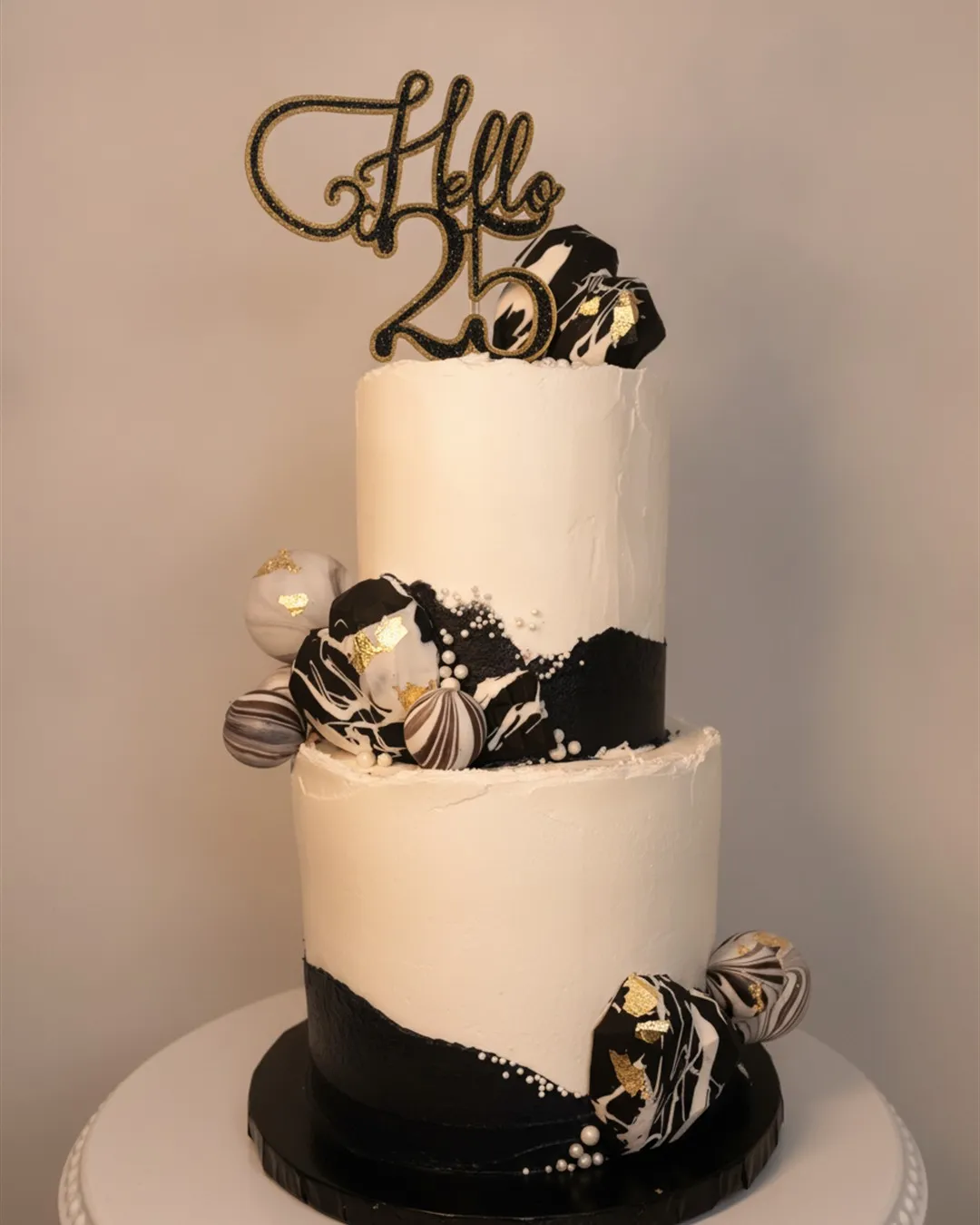 Two-tier cream and black cake with marbled gold spheres and a script milestone topper