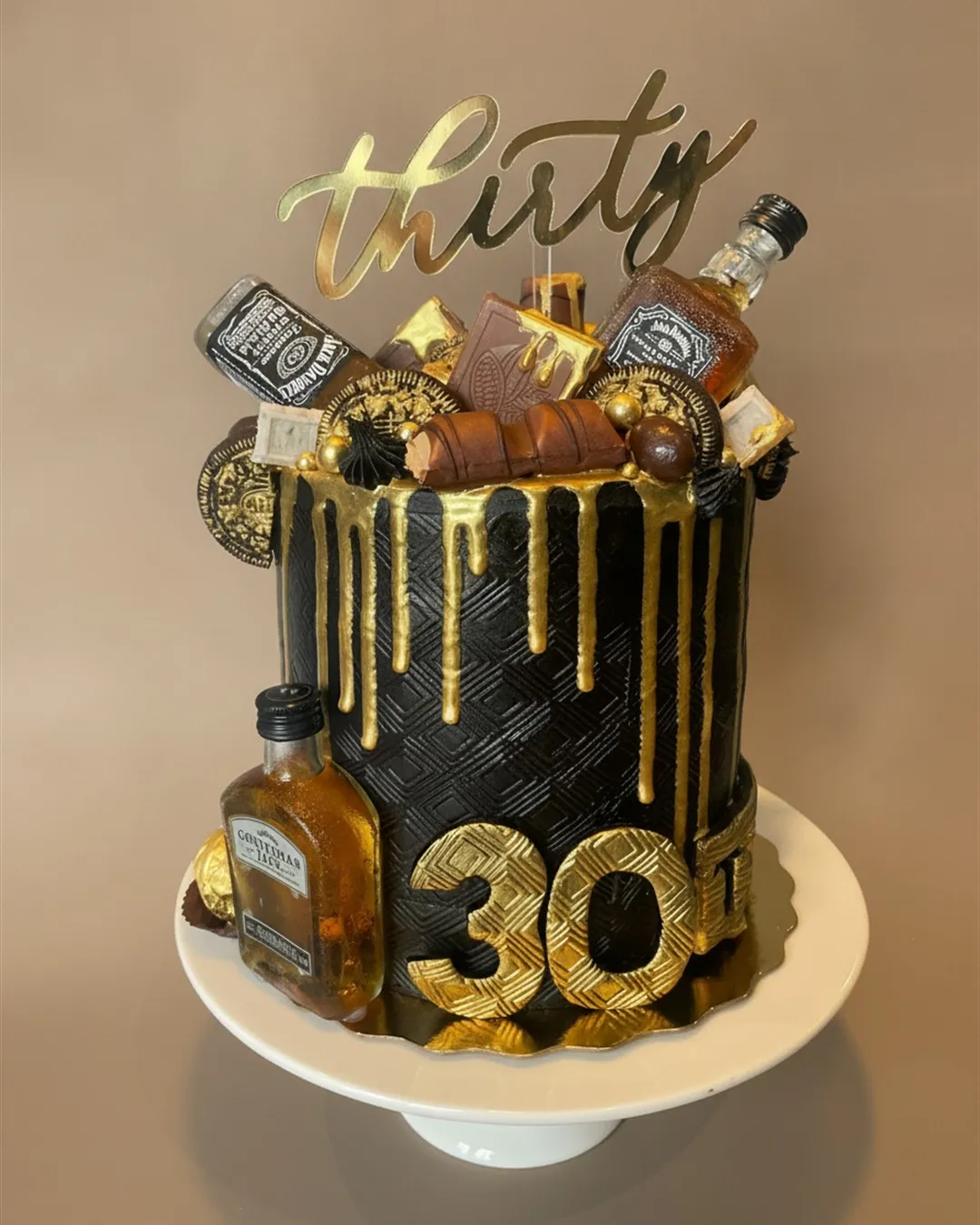 Tall black cake with gold drip, gold number topper and miniature bottle decorations