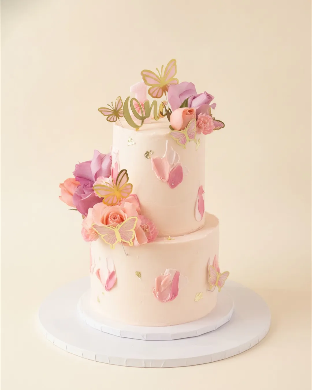 Two-tier blush cake with sugar butterflies, fresh roses and a gold first birthday topper