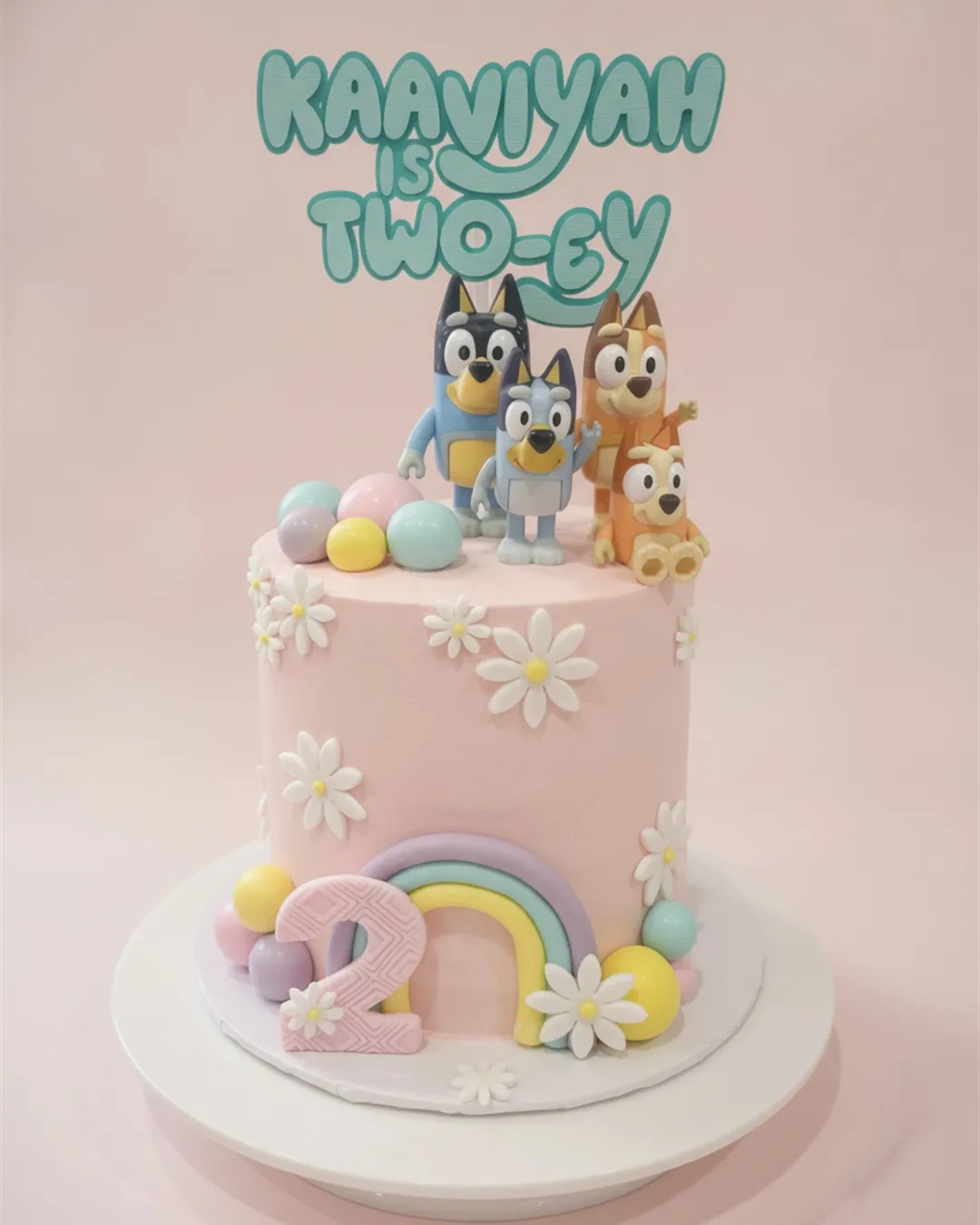 Two-tier pink cake with cartoon dog figures, sugar daisies, a pastel rainbow and a number two