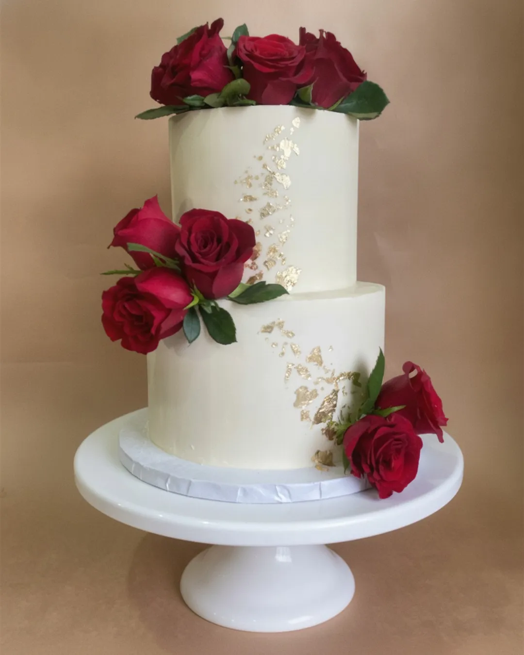 Two-tier white cake with deep red fresh roses and scattered gold leaf detailing