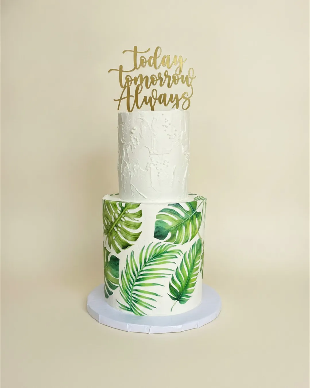 Two-tier white wedding cake with hand-painted green palm leaves and a gold Today Tomorrow Always topper