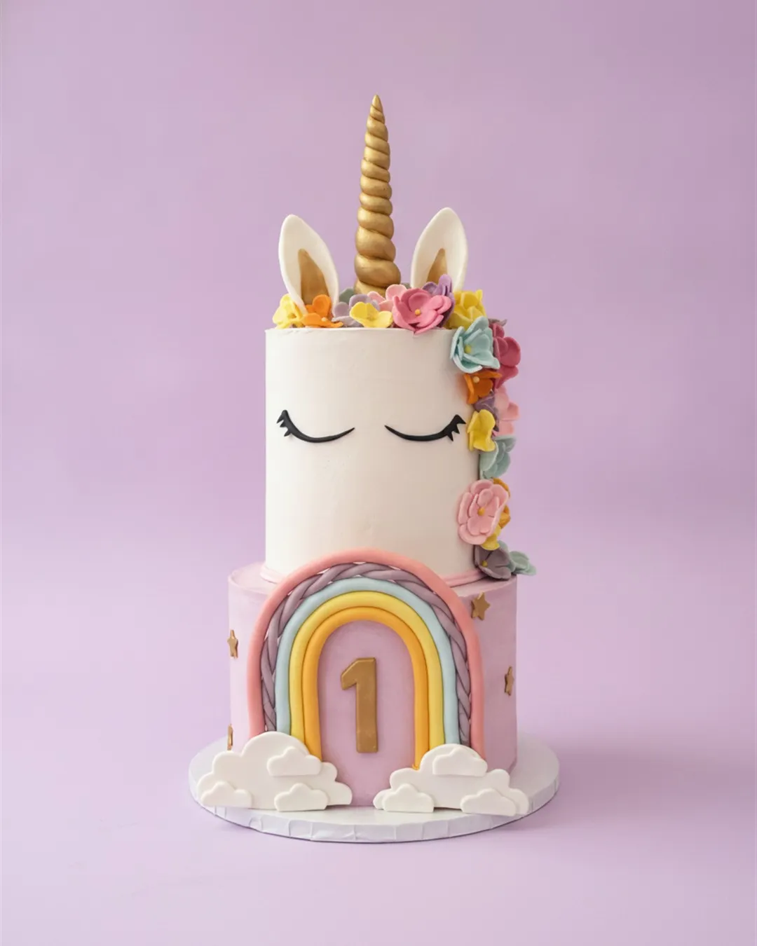Two-tier unicorn cake with a gold horn, sugar flower mane and a pastel rainbow and clouds