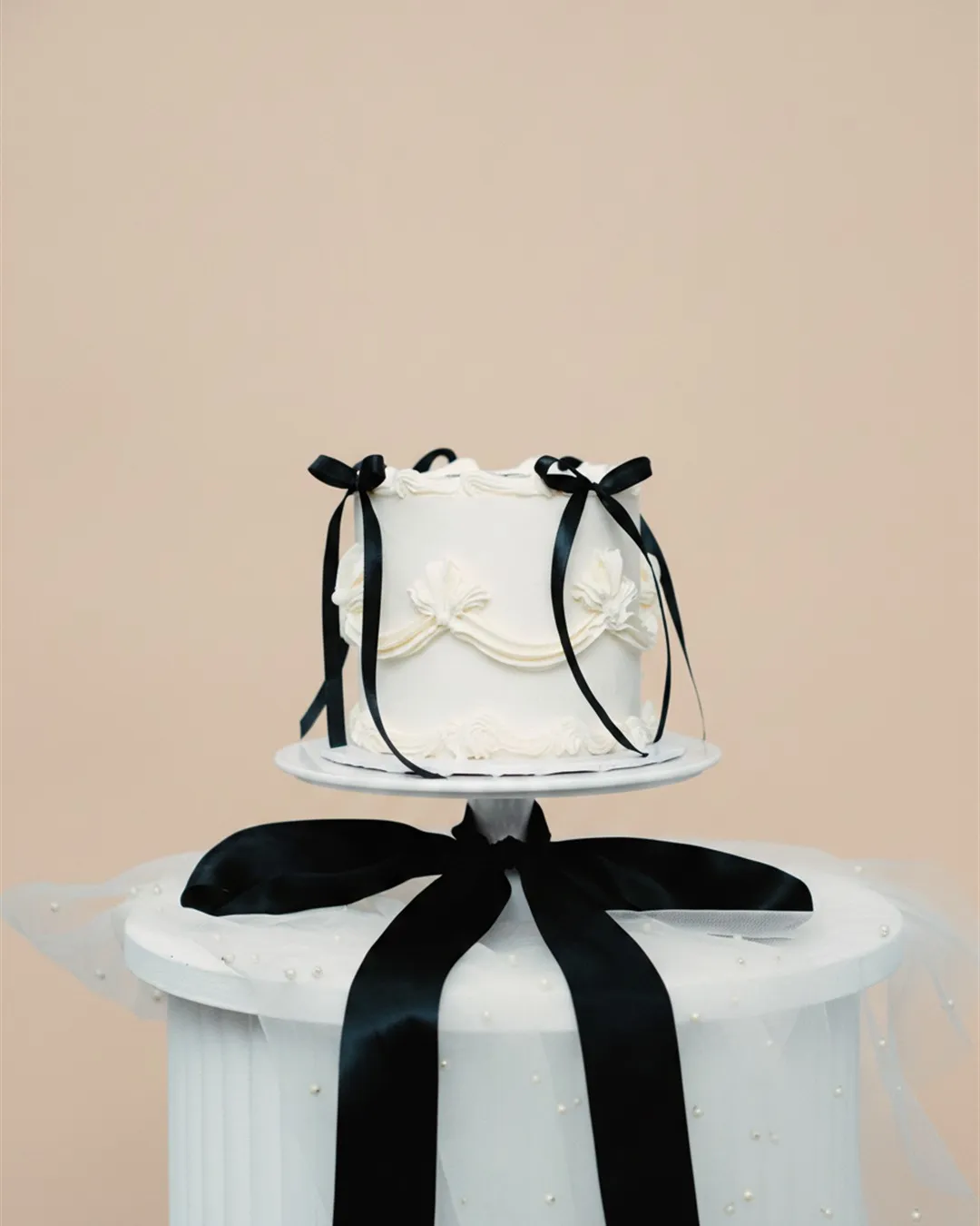 Small white piped cake finished with a black velvet ribbon bow on a pedestal stand