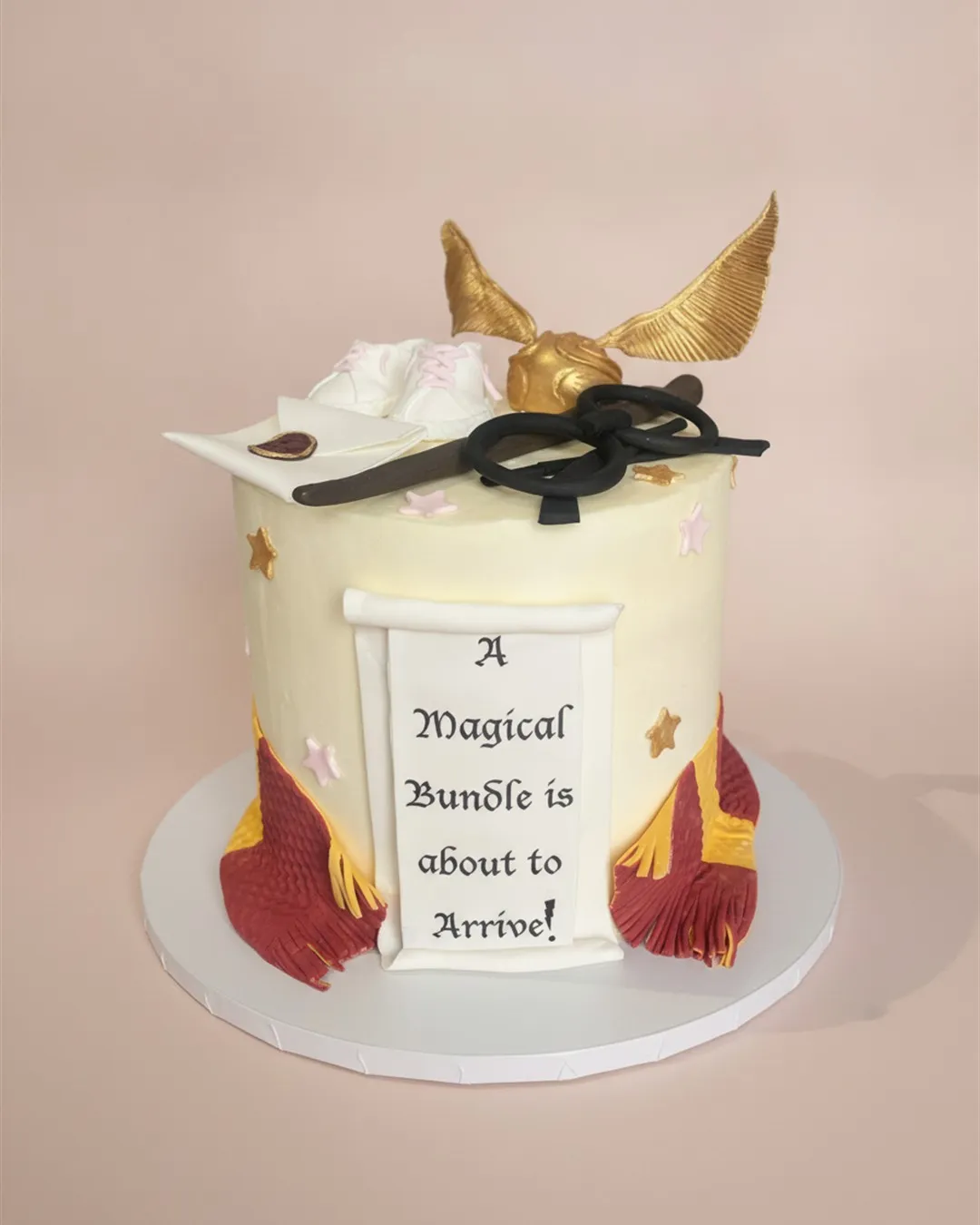Ivory wizard themed baby shower cake with a golden winged key, scroll, scarf and tiny baby shoes